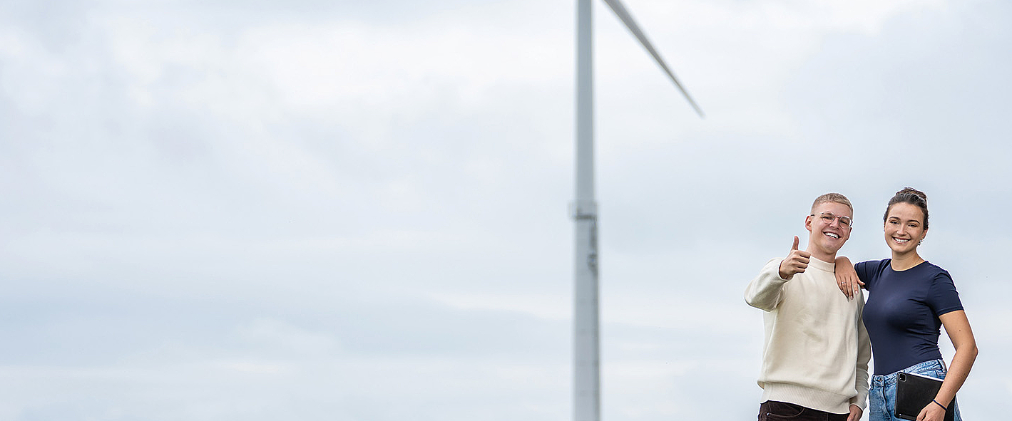 Zwei Studenten, eine Frau und ein Mann, stehen vor einem Windrad und zeigen einen Daumen nach oben in die Kamera