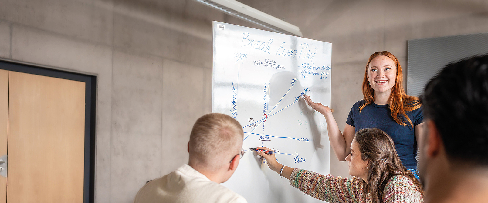 Studentengruppe arbeitet an Whiteboardtafel. Rothaarige Studentin steht am Whiteboard und deutet mit dem Stift auf die Tafel. Sie spricht zu Ihren Kommilitonen