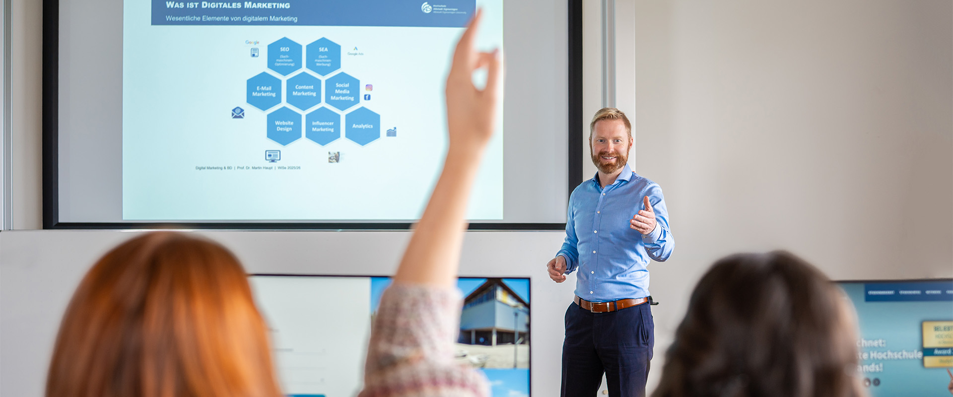Professor Dr. Martin Haupt Ruft Studentin in der Vorlesung auf. Zu sehen ist eine Präsentation an der Wand. Die Studentin ist von hinten, der Professor frontal zu sehen
