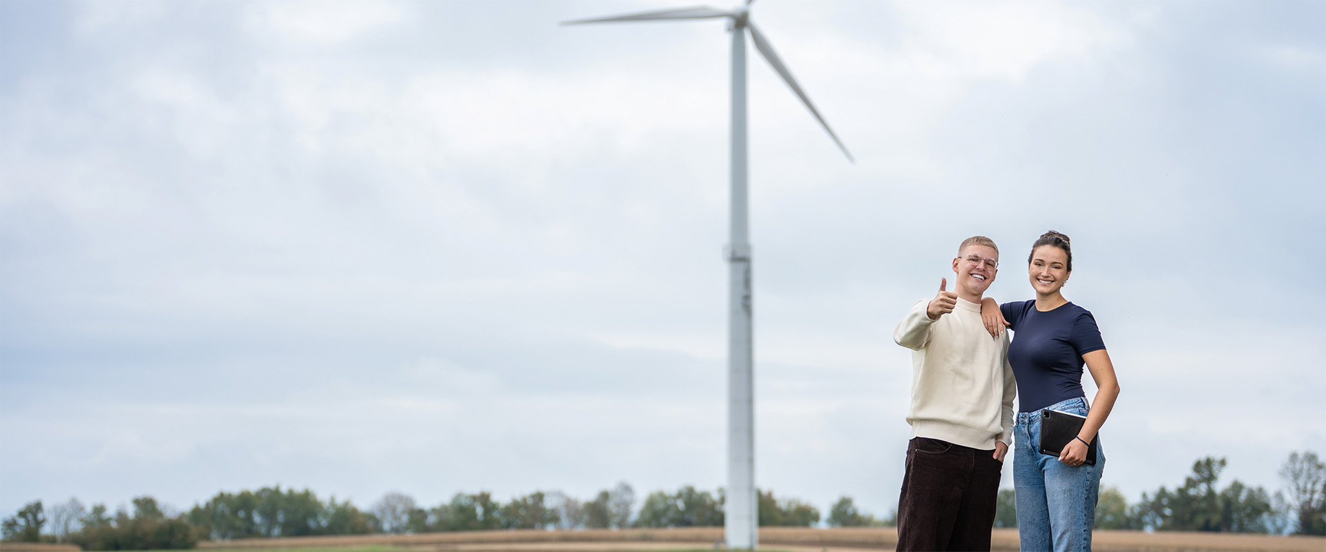 Zwei Studenten, eine Frau und ein Mann, stehen vor einem Windrad und zeigen einen Daumen nach oben in die Kamera